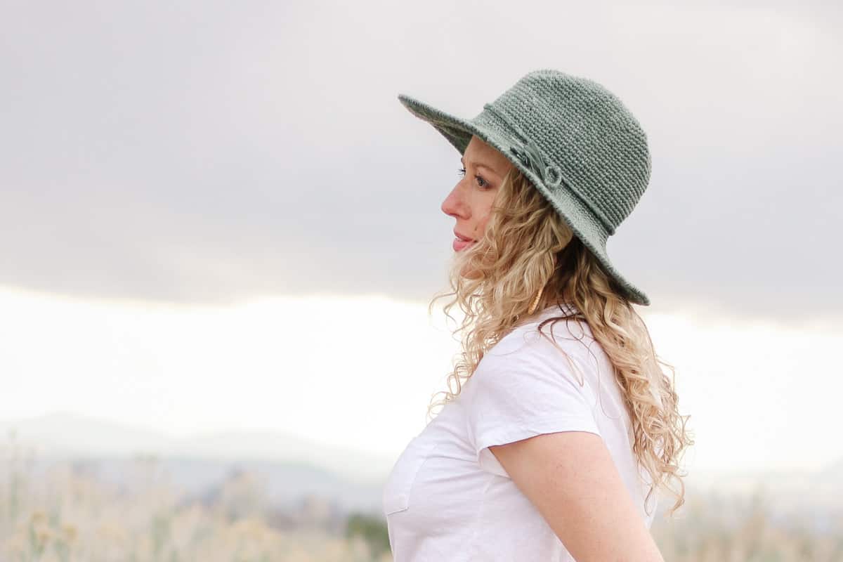 Woman standing in a field wearing a fedora crochet sun hat made with raffia-like yarn.