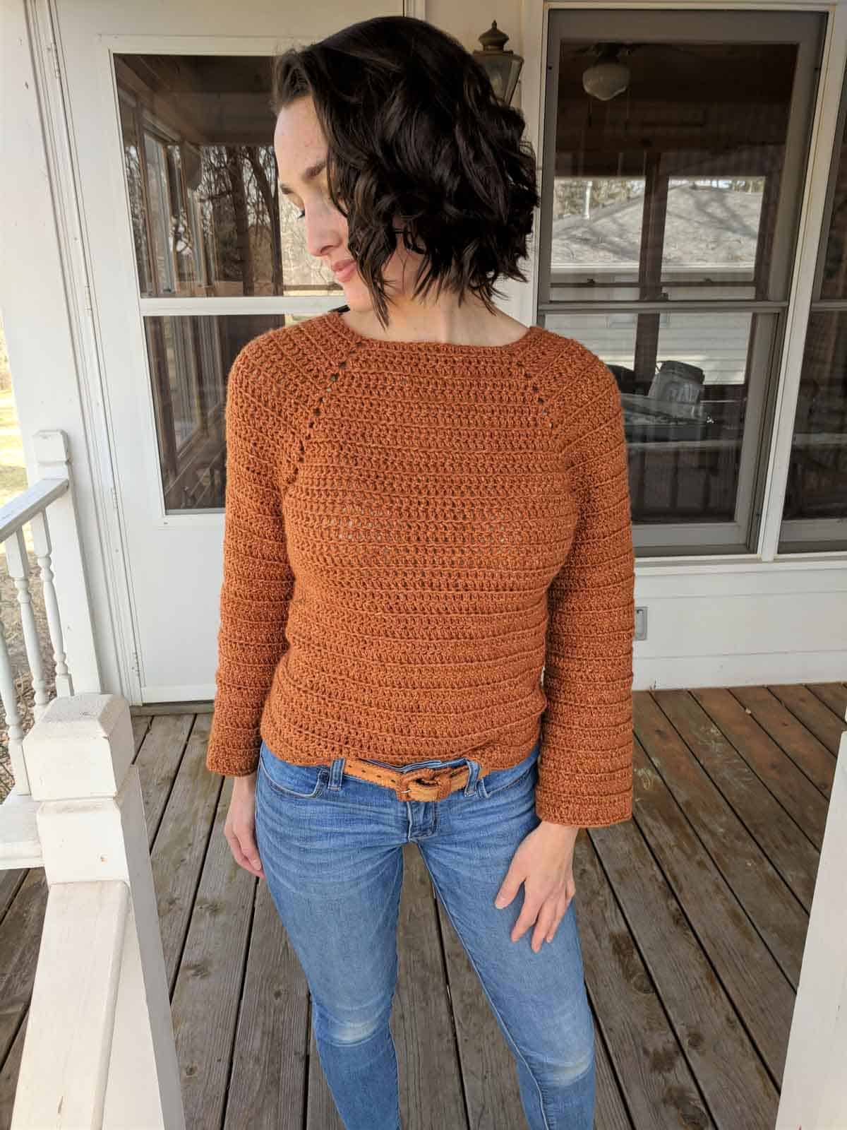 Girl standing outside on front porch of house, wearing blue jeans and a burnt orange crochet sweater.