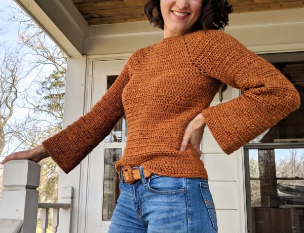 Girl standing outside on front porch of house. She has her hand on her hip and is wearing jeans with a burnt orange crochet sweater with bell sleeves.