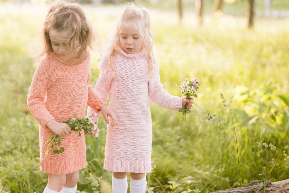 Two girls standing in a field of green grass, wearing crochet sweater dresses and each holding a bouquet of wild flowers.