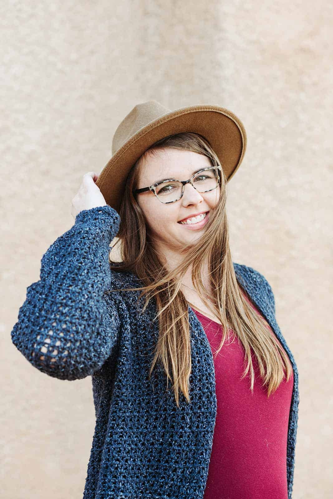 Woman standing in front of a white wall. She has light brown hair and is wearing tortoise shell galsses, a light brown wide-brimmed hat, a red shirt and a lightweight crochet cardigan. She is looking at the camera and smiling while touching her hat. 
