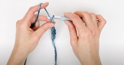 Hands holding yarn and a crochet hook, demonstrating how to create a slip knot and chains for the hat using a crochet hook on a white background.