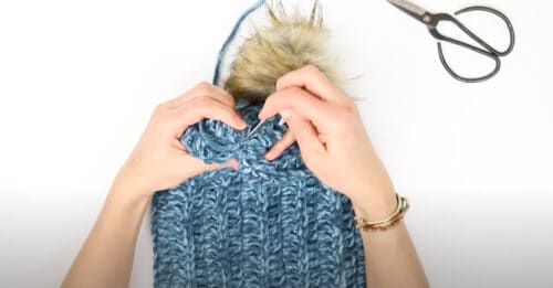 An overhead shot showing how to attach the pom poms on the top part of the hat using a tapestry needle on a white background.
