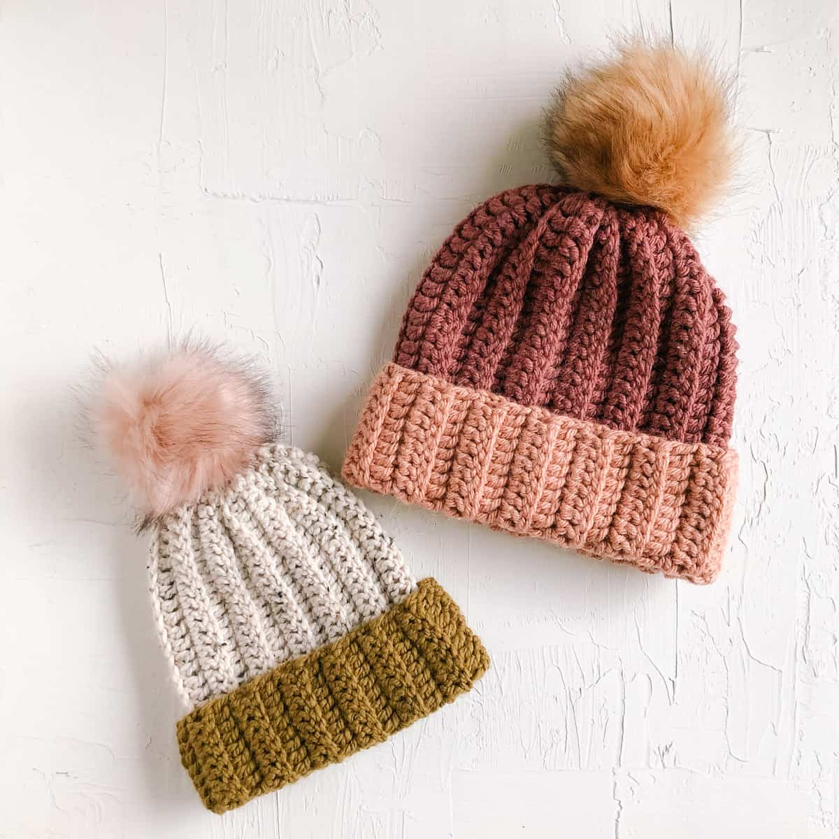 Two simple crochet beanies with faux fur pom poms laying on a white surface.