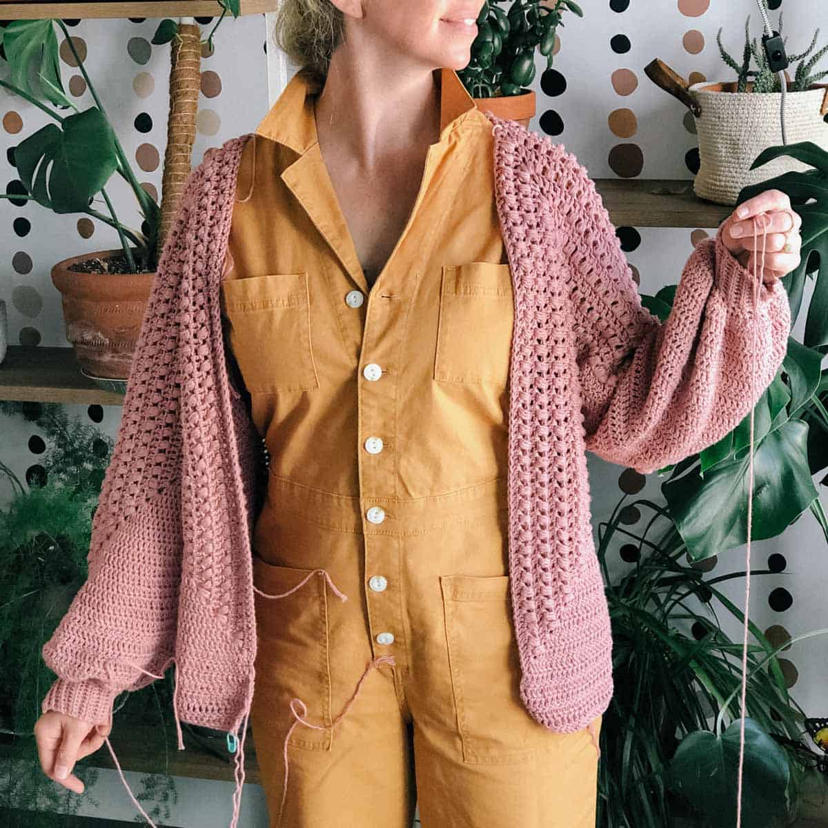 Woman wearing a mustard colored jumpsuit and an in progress crochet cardigan, which still has yarn hanging from it.