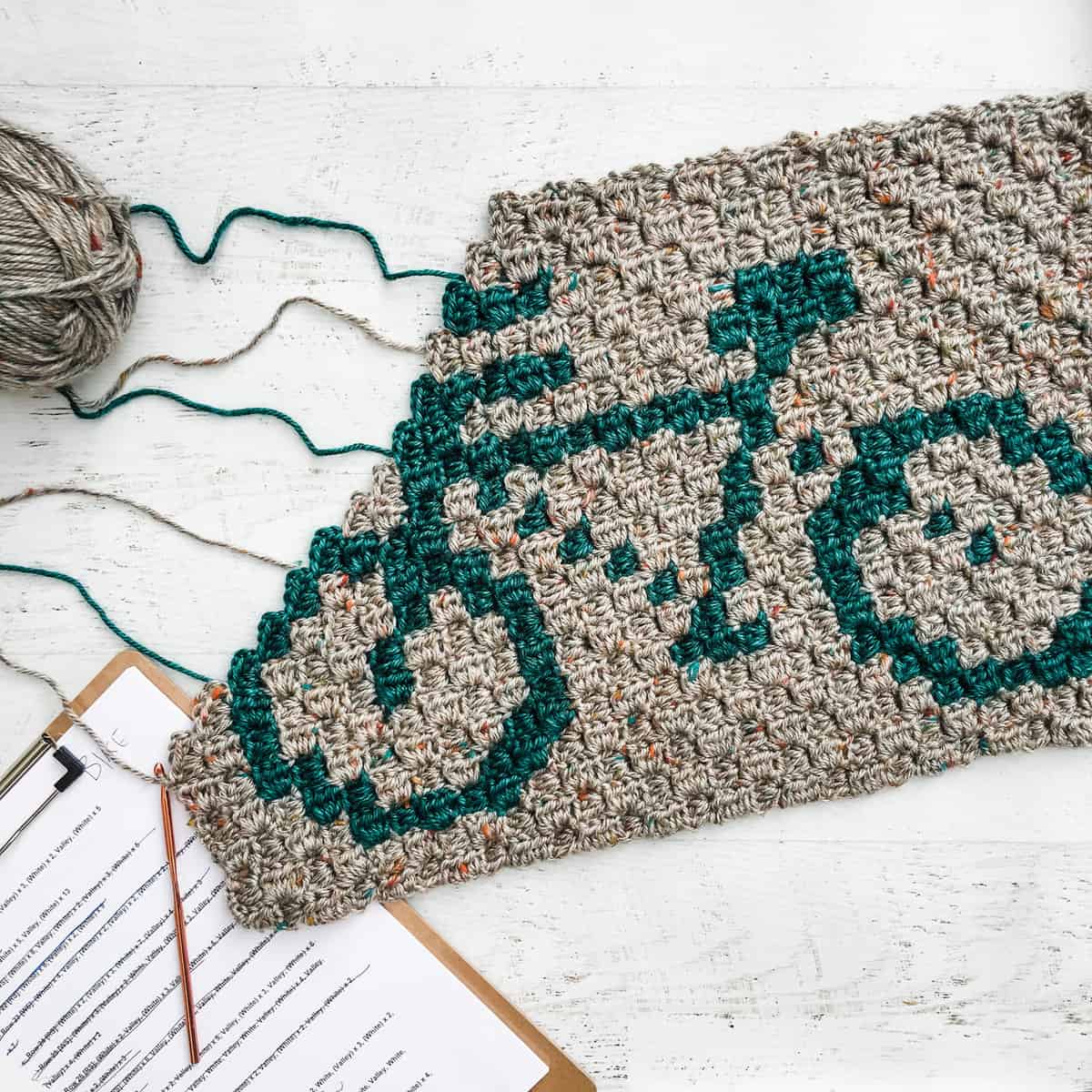 Flat lay of a gray with a green bike pattern C2C blanket, hook, a checklist on the bottom, and a gray yarn on the left side.