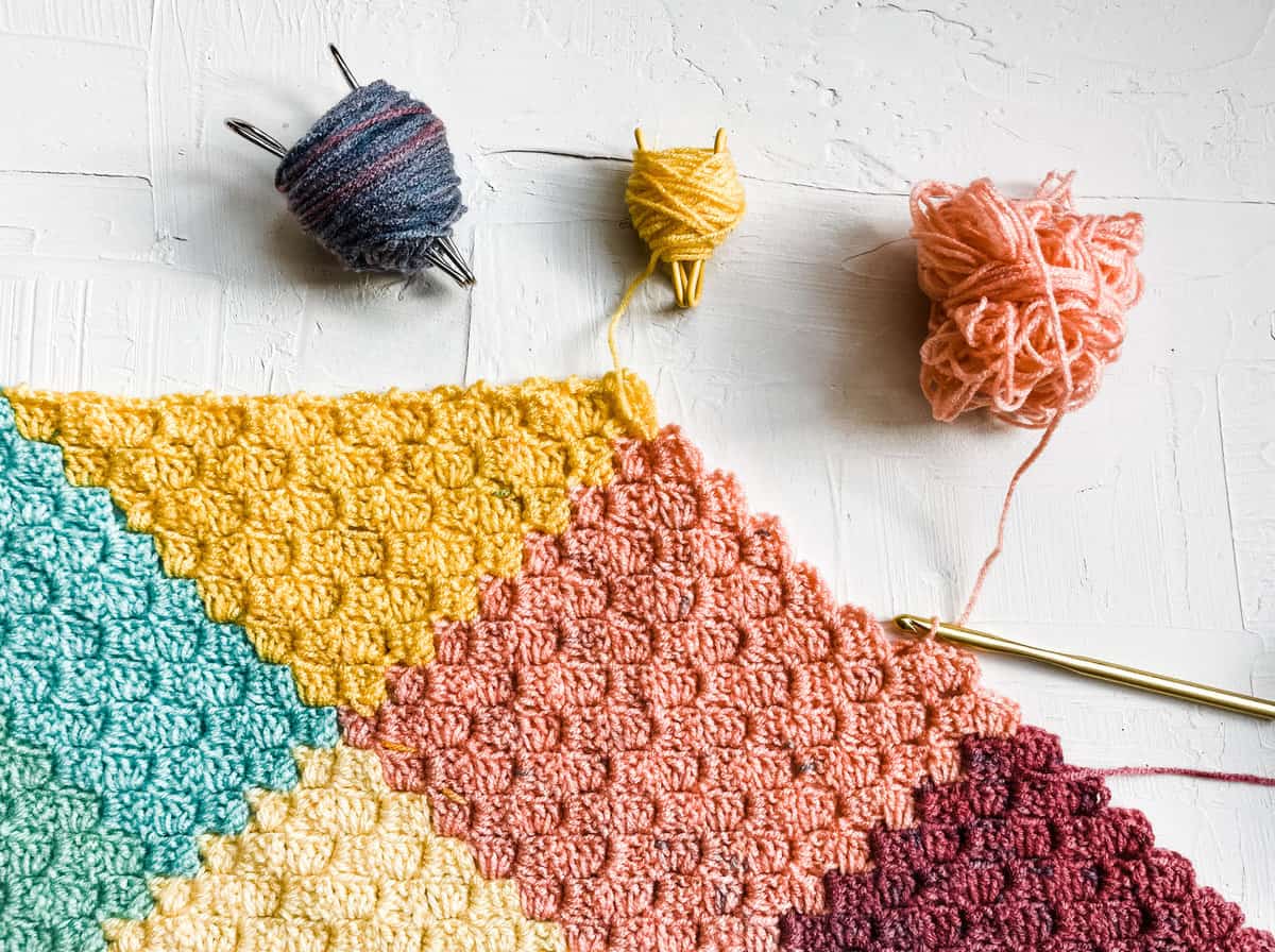 Flat lay of a multi-colored corner-to-corner crochet blanket with three different colored yarns and a crochet hook.