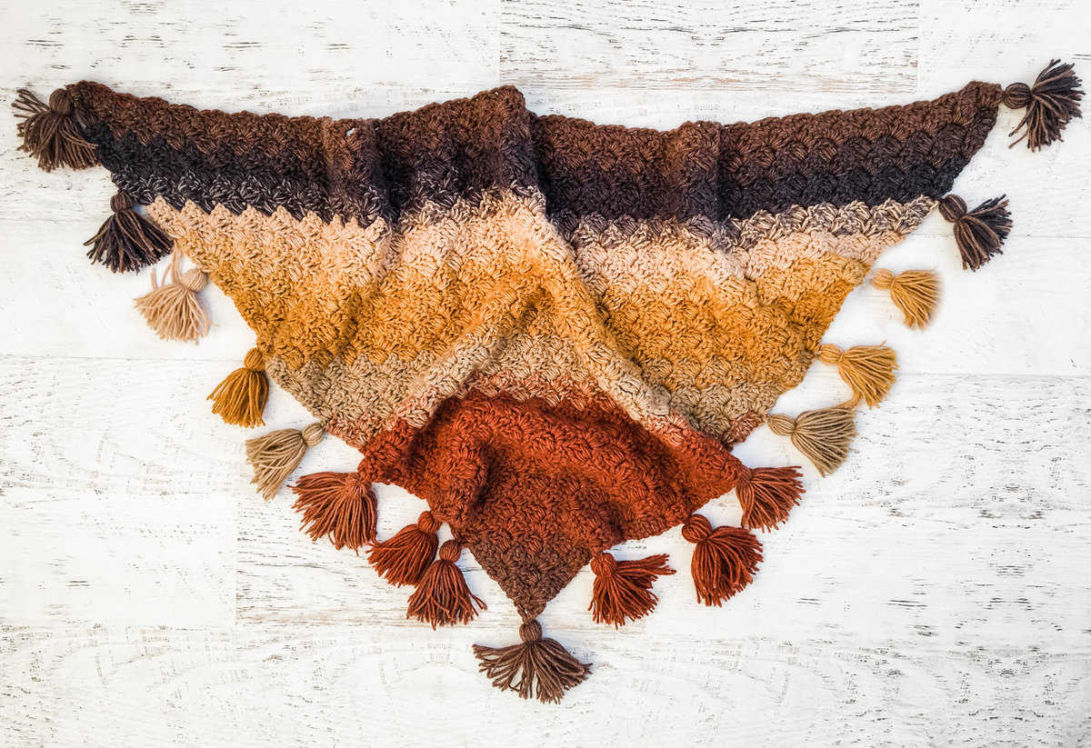 A rustic colored crochet shawl with large tassels laying on a white background.