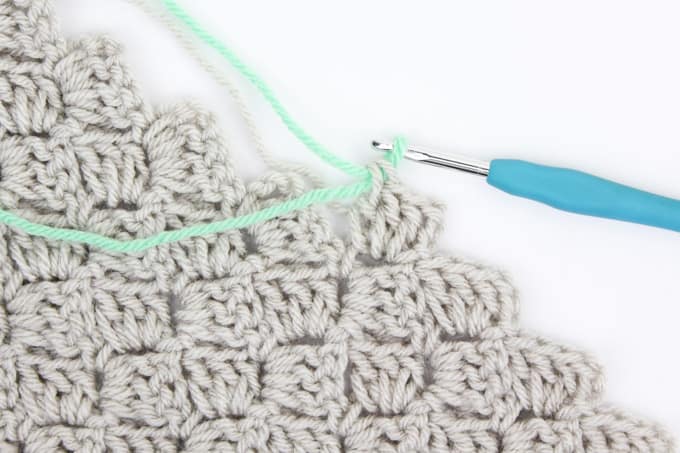 An ivory colored swatch of C2C crocheting in progress. A crochet hook is pulling through a single loop of contrasting teal yarn.