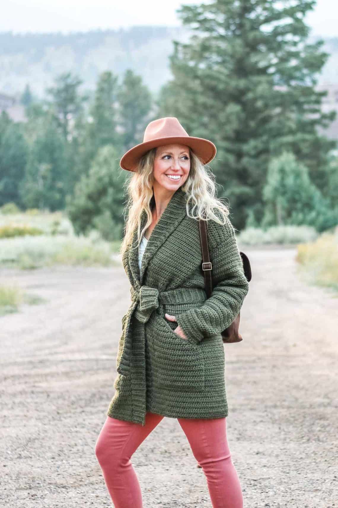 A woman on a dirt road in the mountains wearing a crochet sweater jacket made with Lion Brand Touch of Alpaca yarn in the color "Olive You."