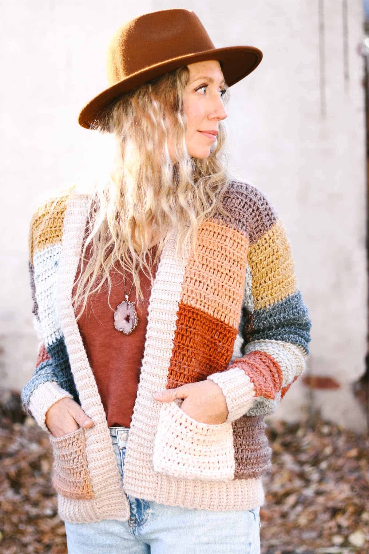 Blonde woman wearing a colorful crochet patchwork sweater with crochet ribbing. Her left hand is in the cardigan pocket.