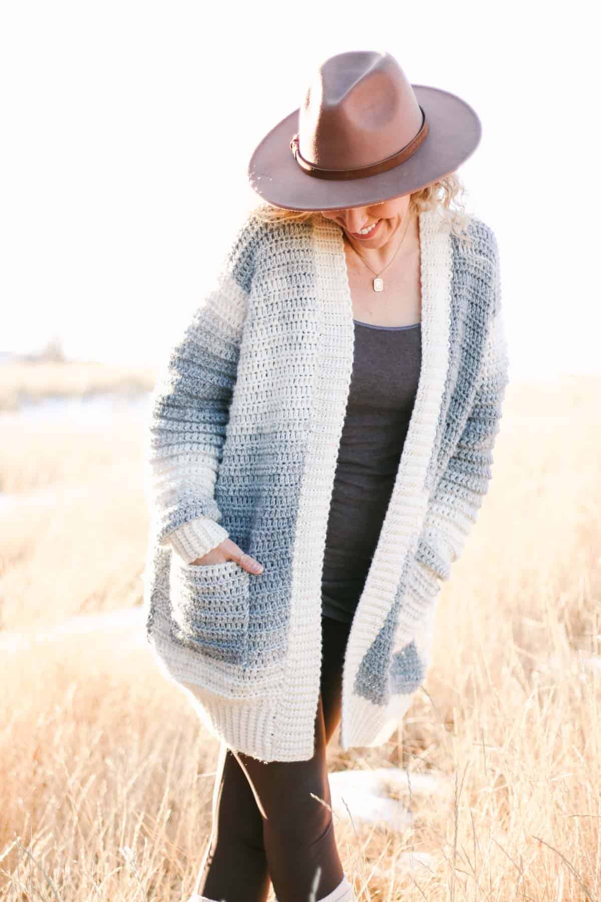 A woman standing in a field of tall grass wearing a neutral toned crochet cardigan with pockets made from Lion Brand Scarfie yarn.