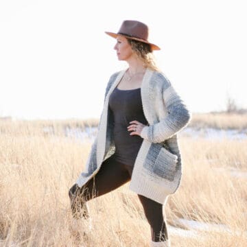 Image of blonde woman standing in grassy field. She is wearing black pants, a black shirt, an ombre crochet cardigan with pockets and a brown hat. Her left hand is on her hip.
