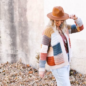 A woman wearing a crochet patchwork cardigan in the aesthetic of Harry Styles. She has one hand raised to her rancher style hat.