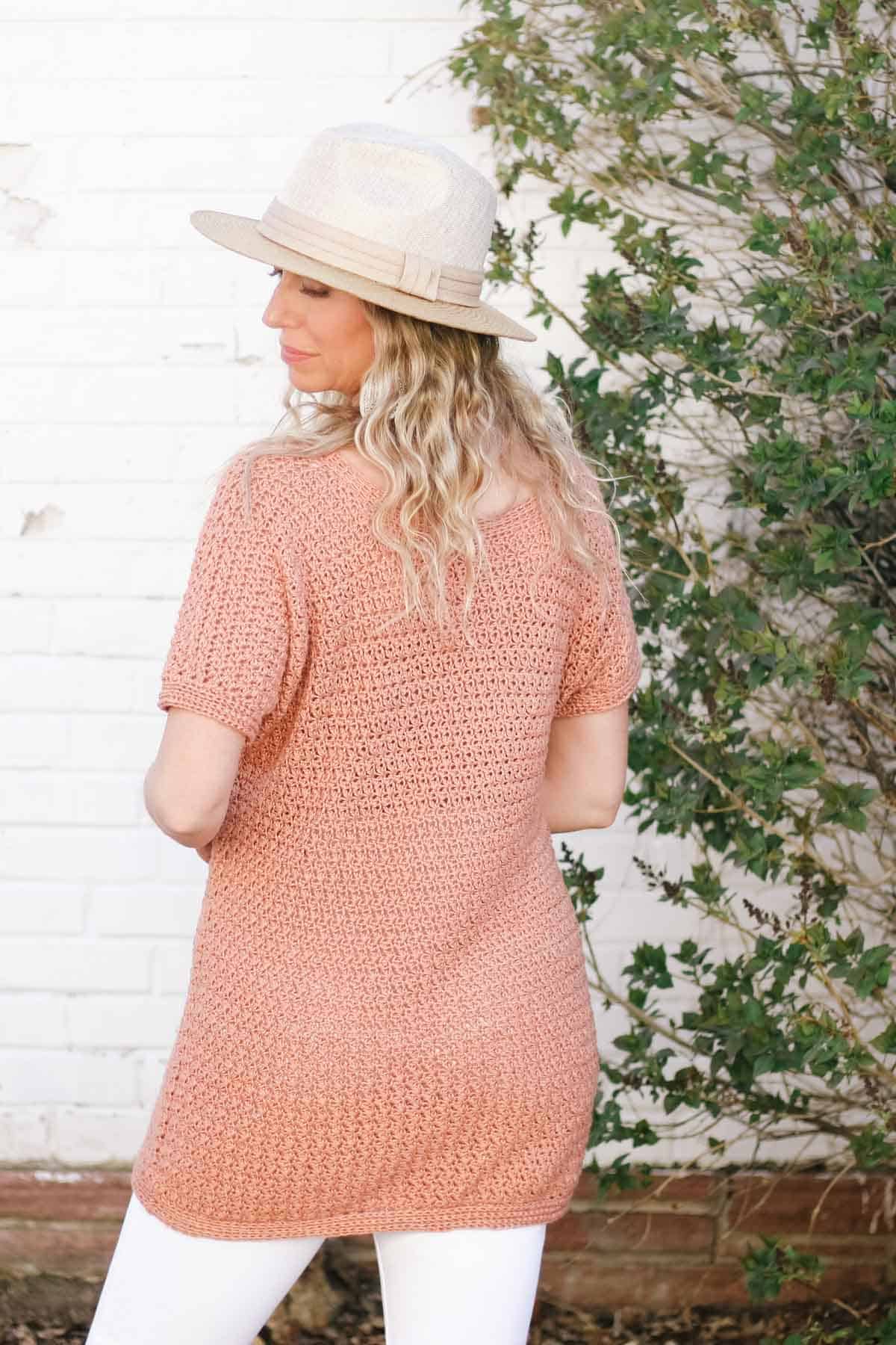 This image shows a woman facing away from the camera standing in front of a white wall with greenery around her. She has blonde hair and is wearing white pants, a light pink colored crochet tunic tee. and a tan hat.