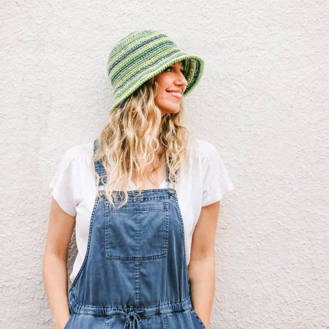 A blonde woman standing against an off white wall wearing a green striped crochet bucket hat made from Lion Brand 24/7 Cotton.