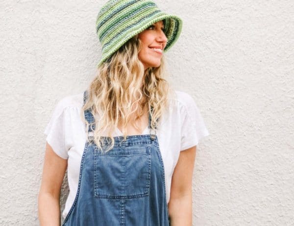 A blonde woman standing against an off white wall wearing a green striped crochet bucket hat made from Lion Brand 24/7 Cotton.