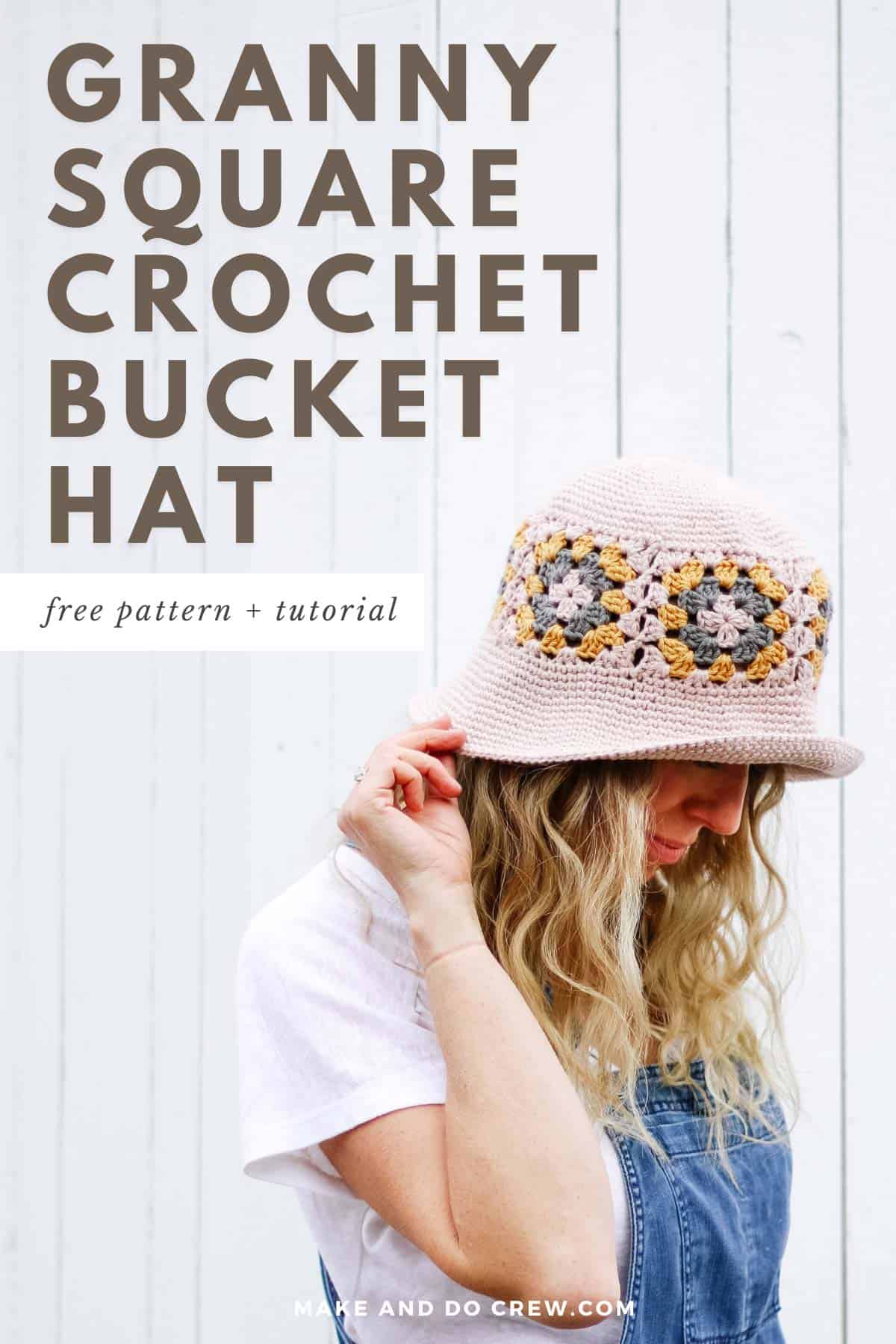 This image shows a woman with blonde hair wearing overalls and a white shirt. She is wearing a crochet bucket hat made out of granny squares. She has her right hand touching the brim of the hat and she is looking down at the ground.