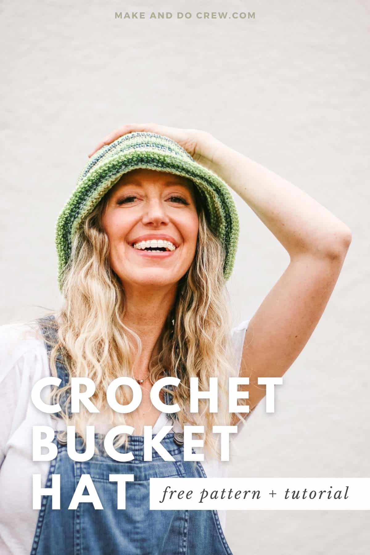 Awoman with blond hair looking at the camera and laughing. She is wearing a green striped crochet bucket hat and her hand is on her head.