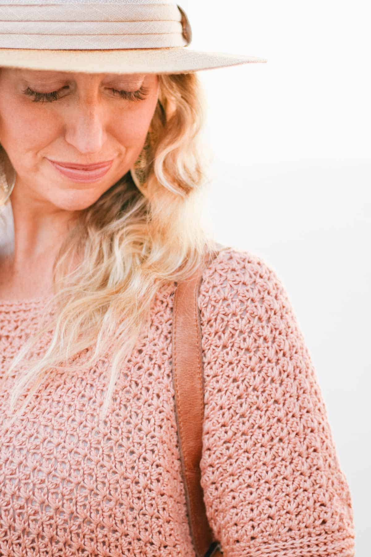 This image shows a woman facing the camera, looking down. It is a close up of her face and shoulder. She is wearing a pink crochet tunic and has blonde hair.