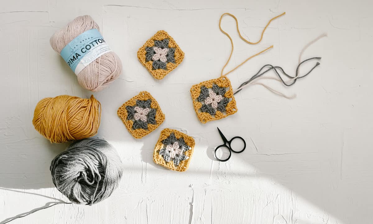 This image shows skeins of Lion Brand Pima cotton yarn, four crochet granny squares and a pair of tiny scissors.