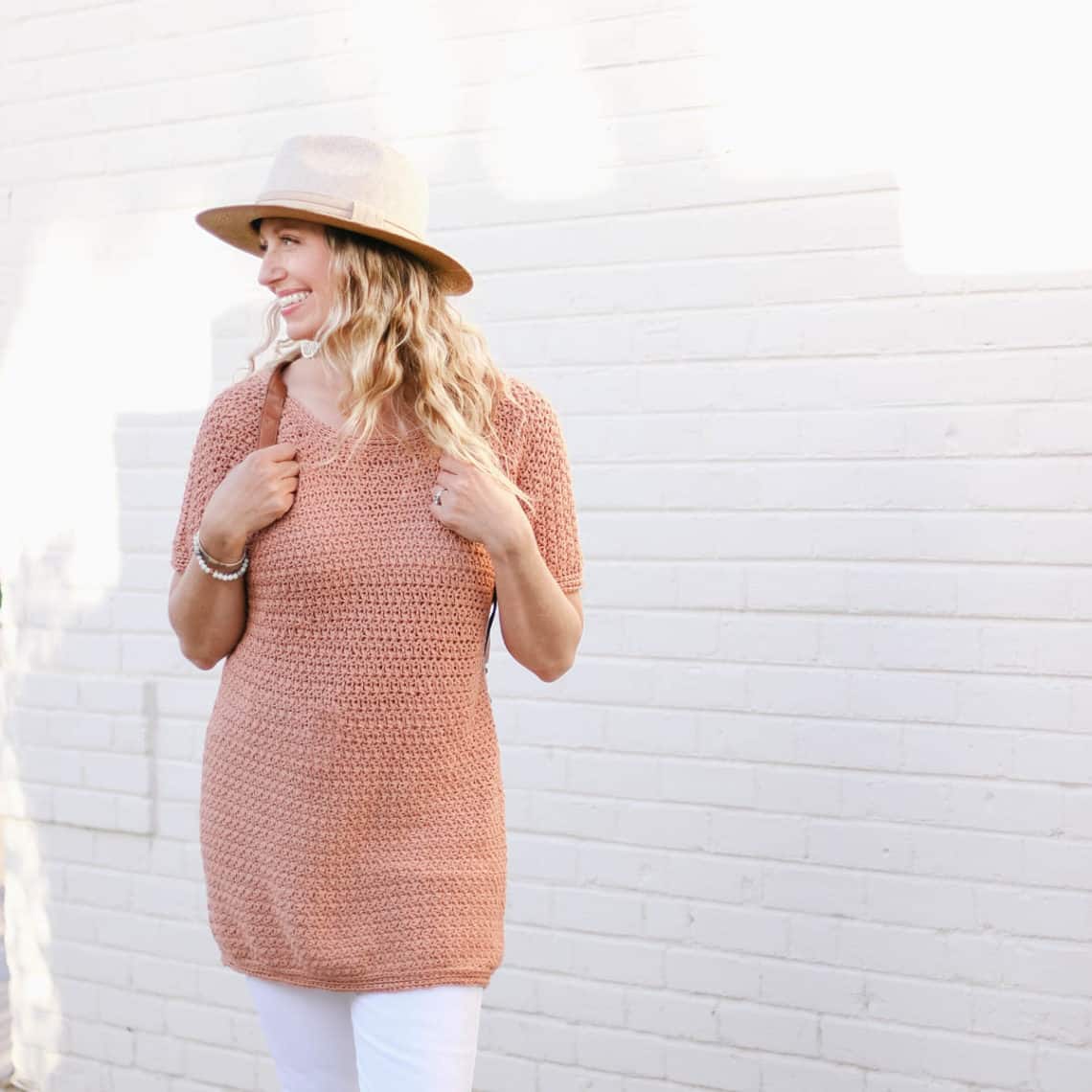 This image shows a woman standing with her arms crossed in front of a white brick wall. Her body is facing the camera and she is looking over her shoulder. She is wearing a mauve colored crochet tunic, white pants and a tan hat.