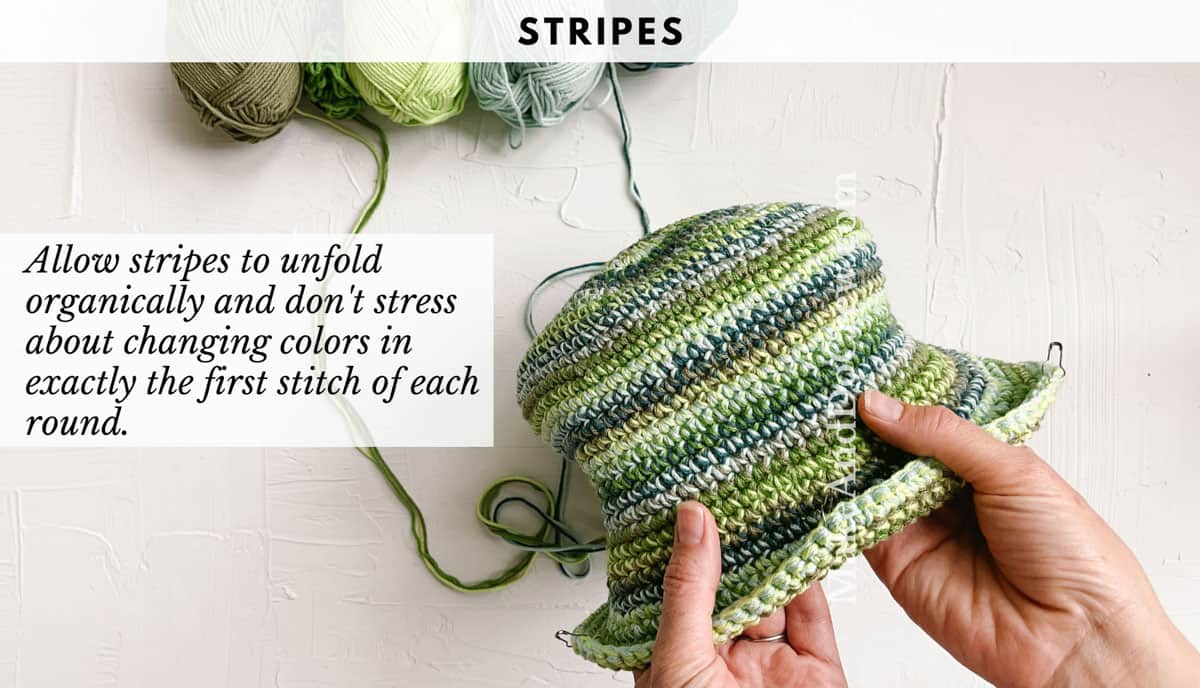 This image shows a green striped crochet bucket hat. A woman's hands are holding the hat and there are balls of yarn attached.