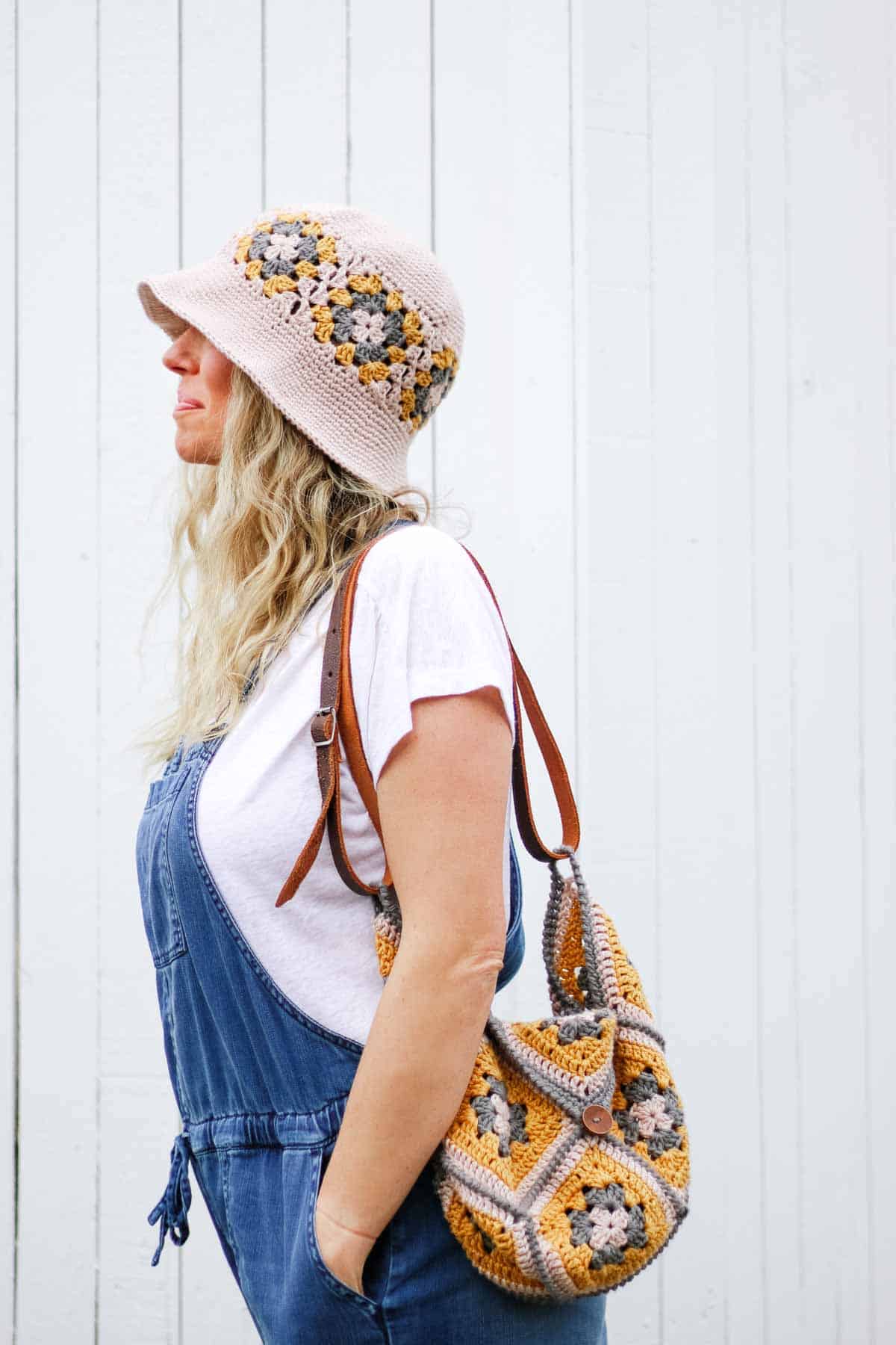 A woman with blonde hair wearing overalls, a white tee shirt, a crochet granny square hat and a crochet granny square purse with a leather strap. She is looking and facing left with her hand in her pocket.