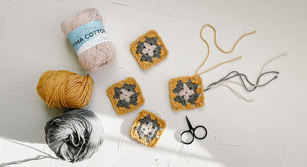 This overhead image shows four in-progress crochet granny squares and three balls of Lion Brand Pima Cotton yarn in muted colors.