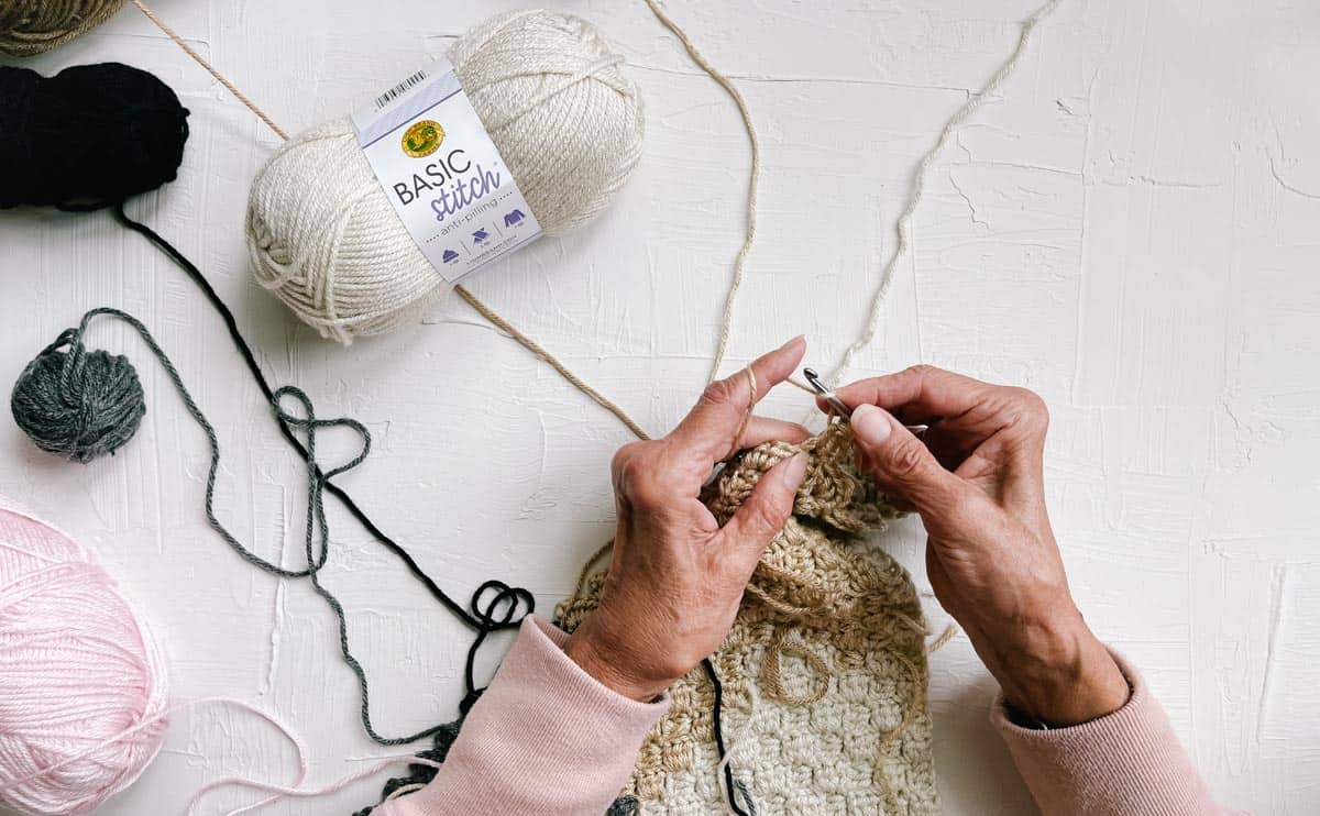 An overhead shot of a woman's hands crocheting using a basic stitch yarn that is scattered in the white background. 