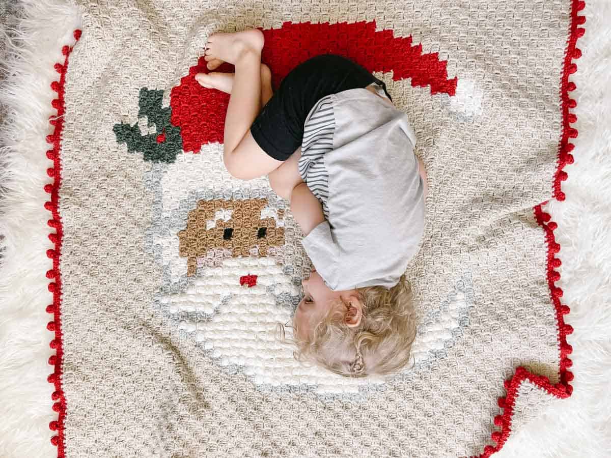 This overhead image shows a toddler with blonde hair wearing a gray shirt and black shorts. He is curled up on a crochet blanket that features a Santa Clause face.
