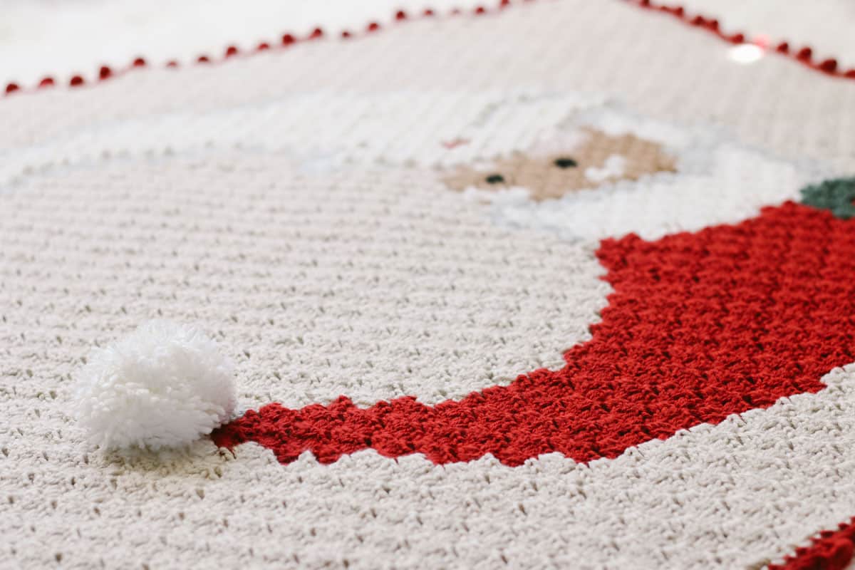 This angled image shows a close up photo of a white pom pom attached to a red santa hat on a crochet blanket.