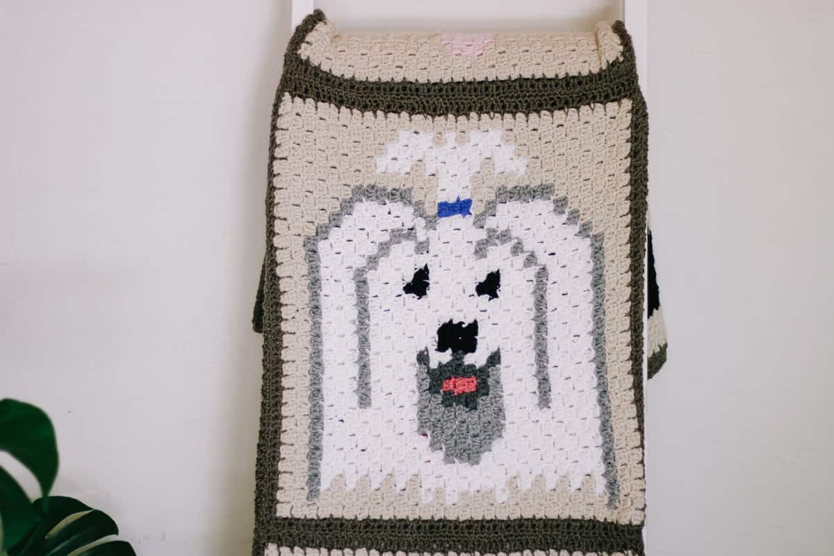 This image shows a white and gray havanese dog crochet blanket square. The blanket is folder onto a blanket ladder and is in front of a white wall.