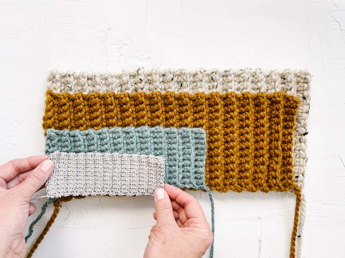 A series of four crochet rectangles made of scrap yarn in different sizes stacked on top of each other. A woman's hands are reaching into the frame to grab one rectangle.