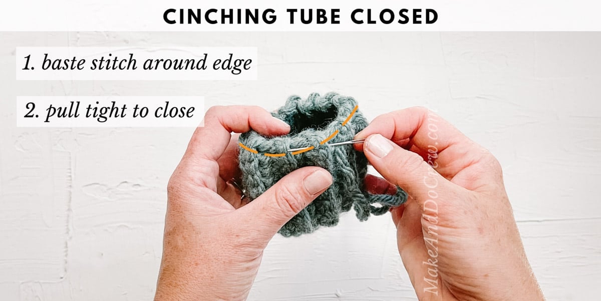 A woman's hands holding a tapestry needle and a crochet rectangle folded into a tube shape. She is demonstrating how to cinch the tube into a pumpkin shape.