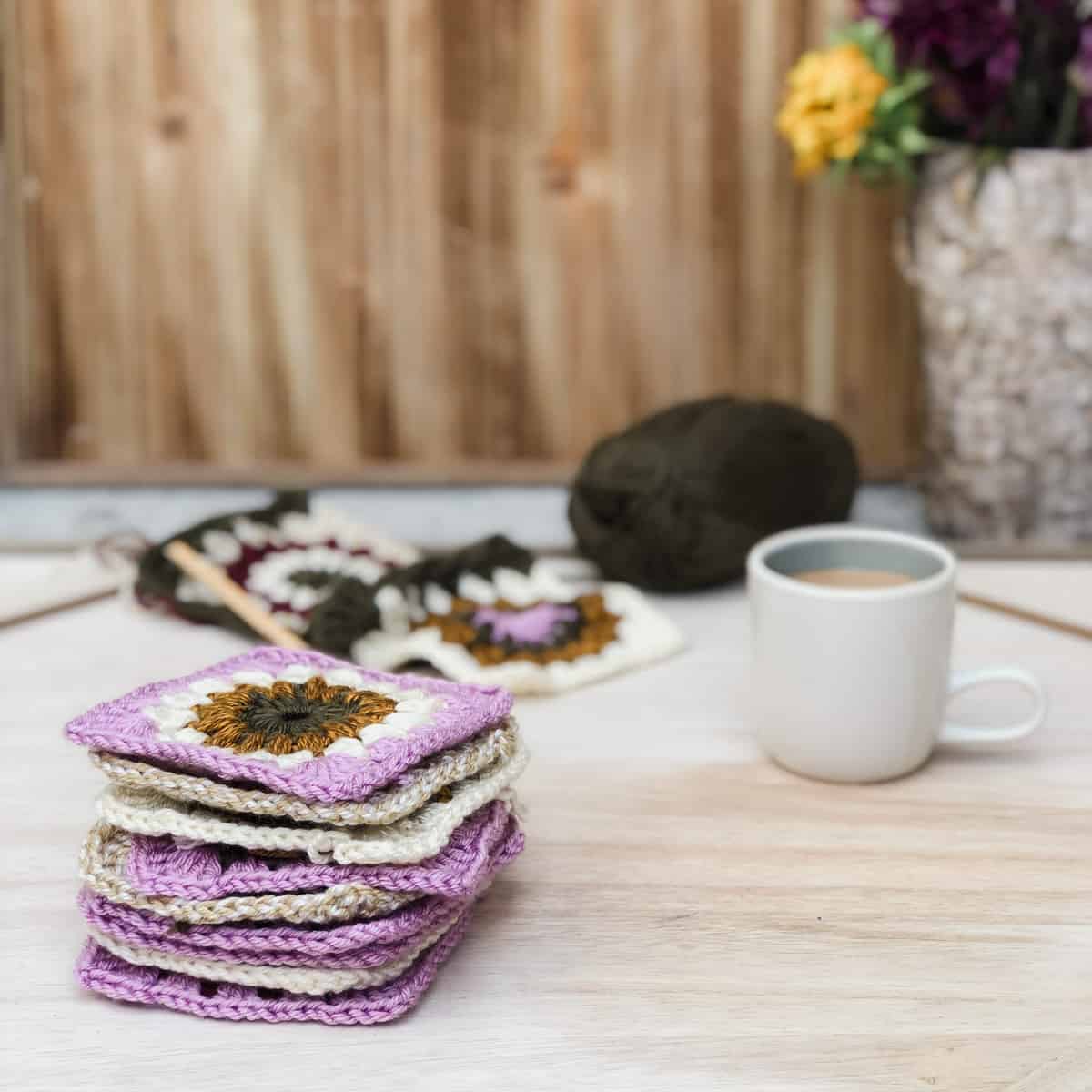 On top of a wooden table are a stack of crochet granny squares, a white mag, an in-progress joined granny squares, and yarn.