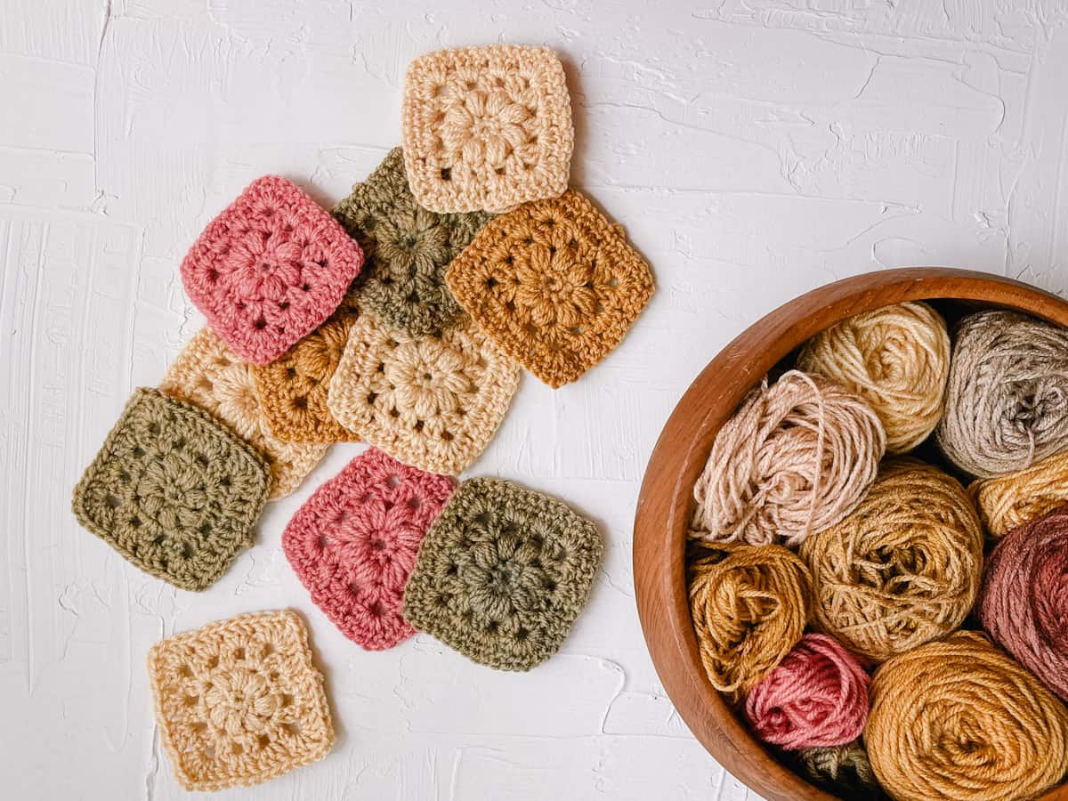Flat lay of eleven multi-colored granny squares next to a wooden bowl filled with balls of yarn on a white background.