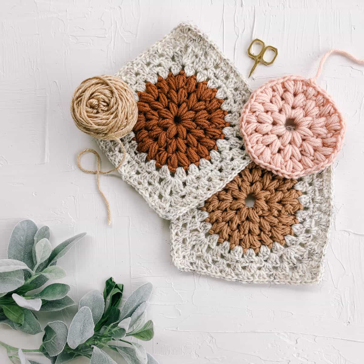 A flat lay of three Dahlia scrapagan granny squares is in-progress with yarn, scissors, and a plant on a white background. 