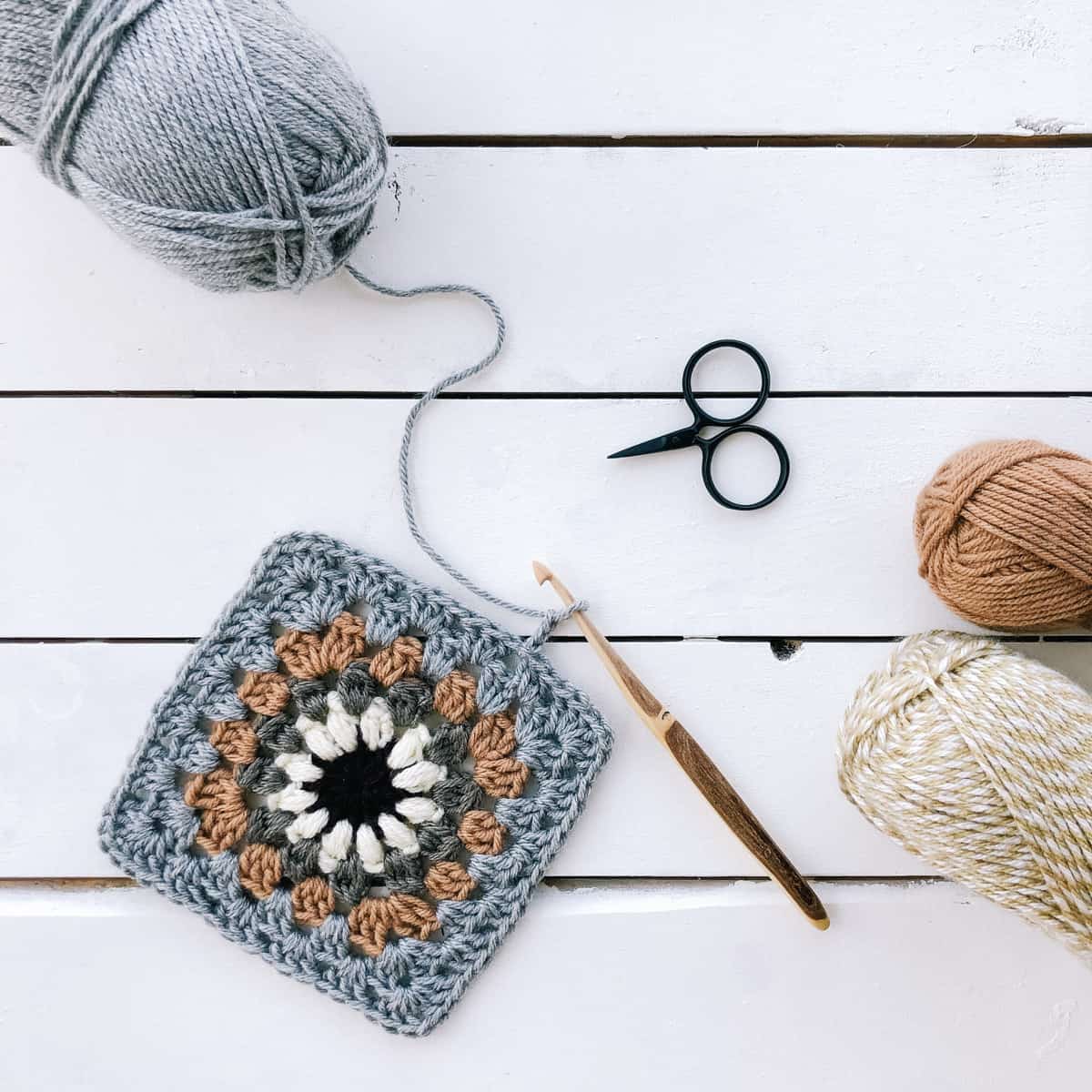 Flat lay of an in-progress granny square crochet with a wooden hook, three yarns, and scissors on top of a white table.