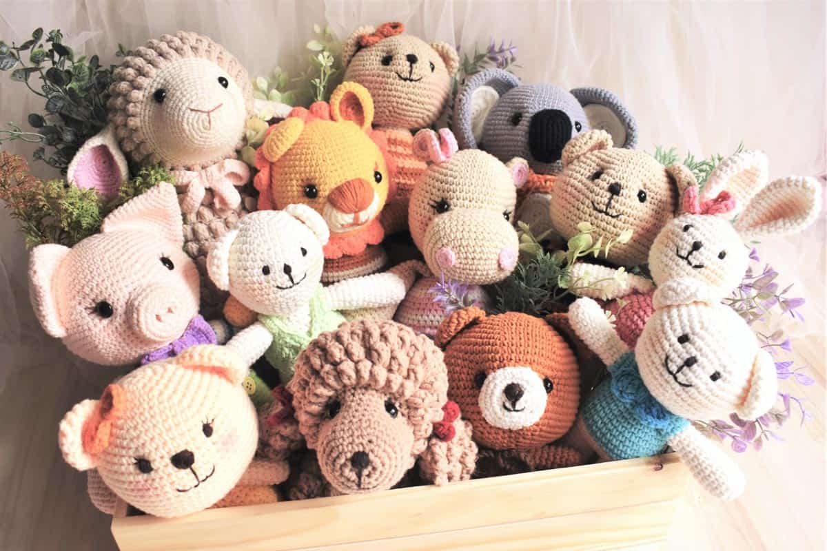 A collection of 13 different crochet animal amigurumi dolls on a wooden box with faux plants.