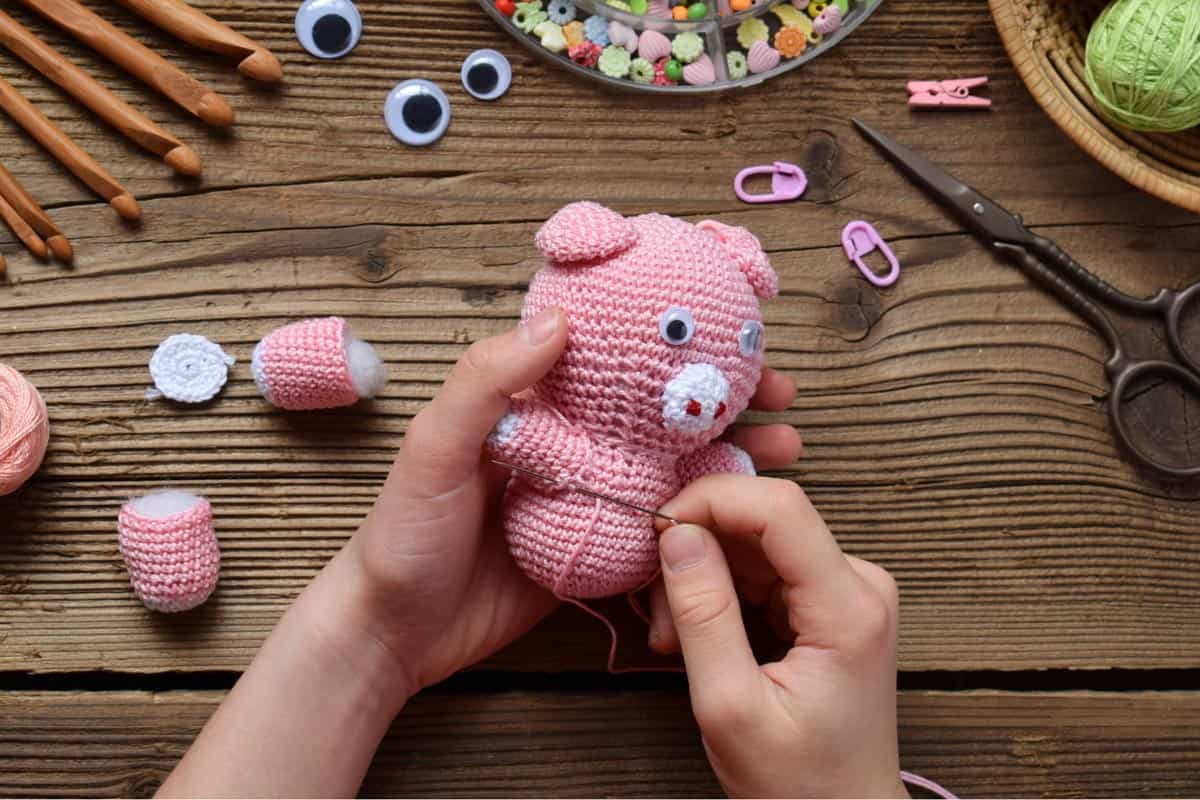 An amigurumi pink bear with a needle to crochet the bear's arms and legs on top of a wooden table.