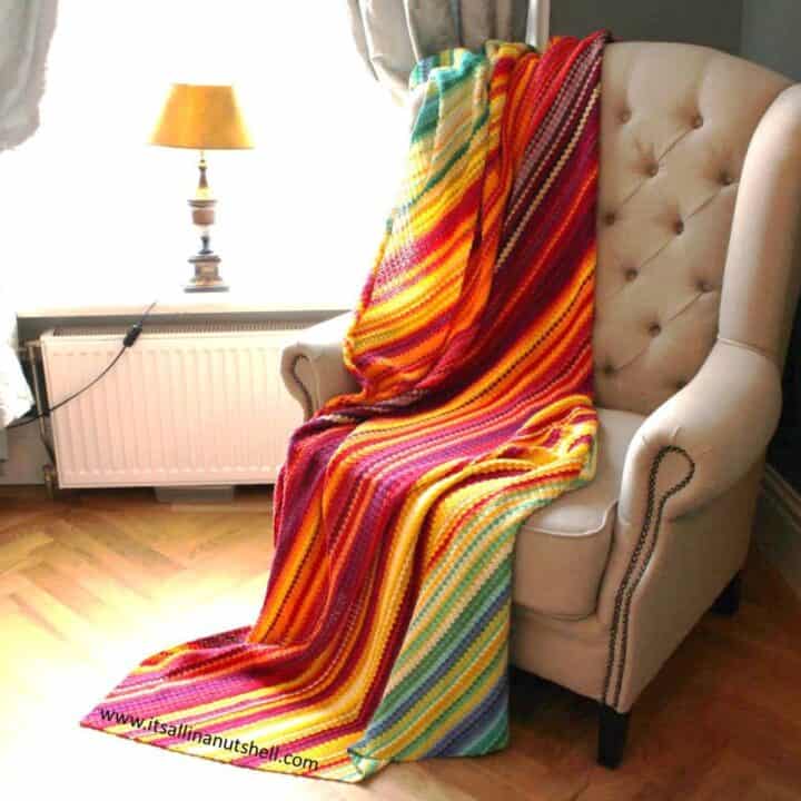 A corner-to-corner crochet temperature blanket draped on a chair beside the window with a table and a lamp.