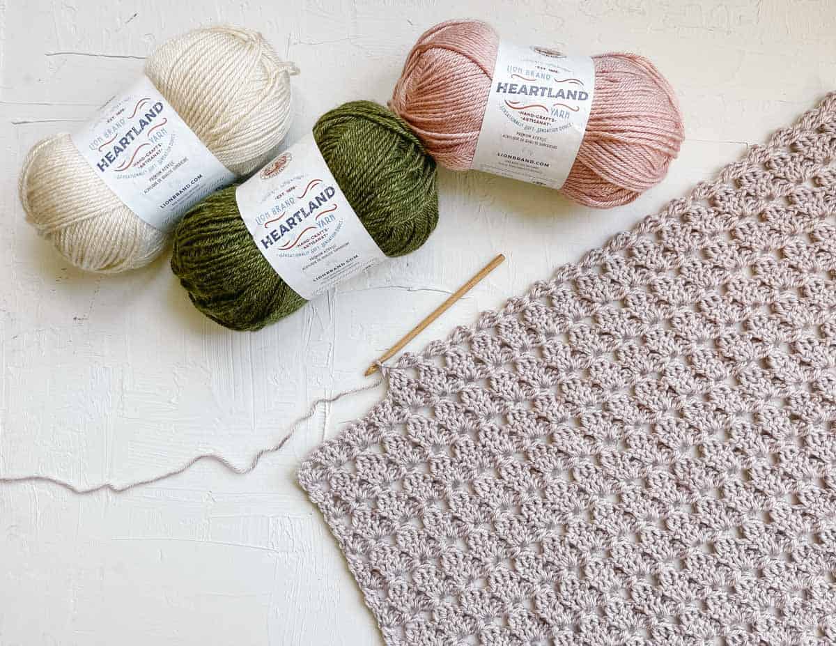 Three Lion Brand Heartland yarn skeins with an in-progress lace pattern crochet scarf and a wooden hook.