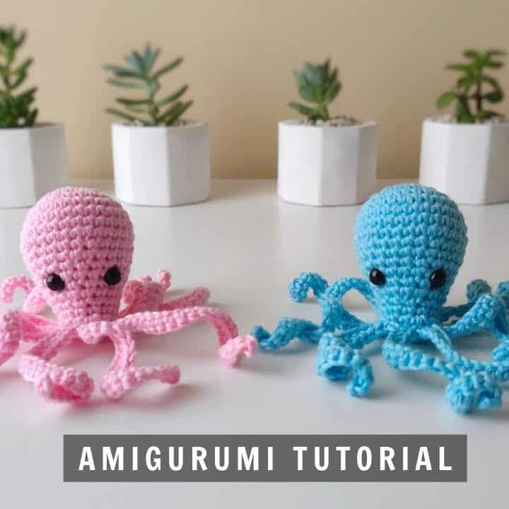 A pink and blue colored octopus amigurumi with black eyes on a table with plants at the back.