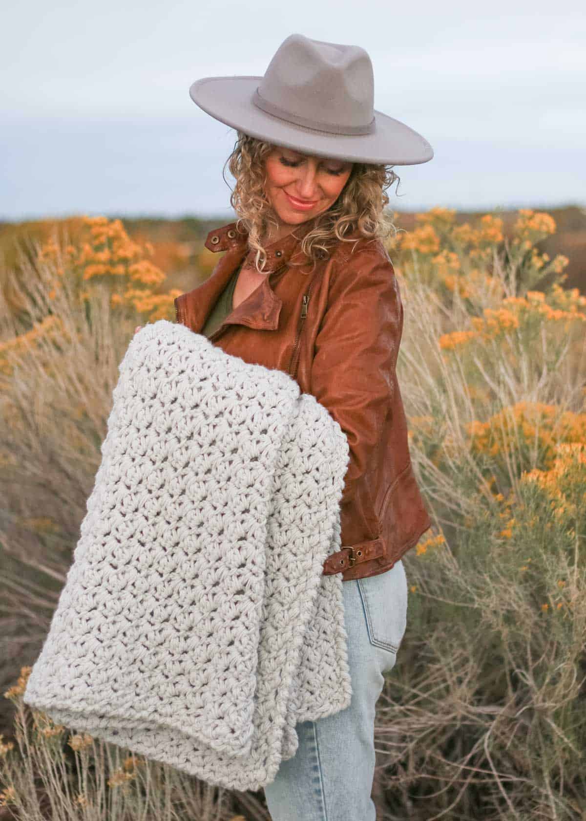 A woman looking down, standing on a field, wearing a fedora hat, a leather jacket, and holding a chunky crochet blanket.