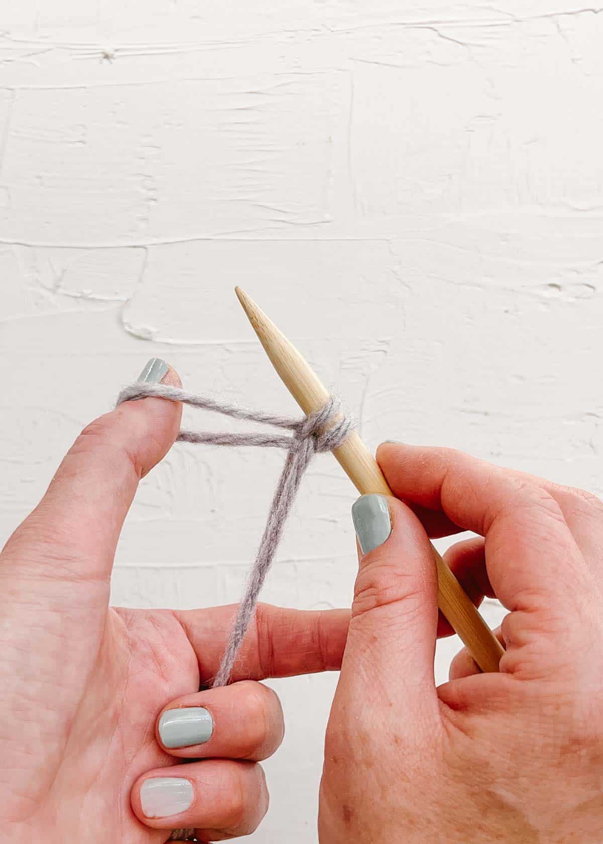 Wooden knitting needle dropping loop of purple yarn in sixth step of casting on.