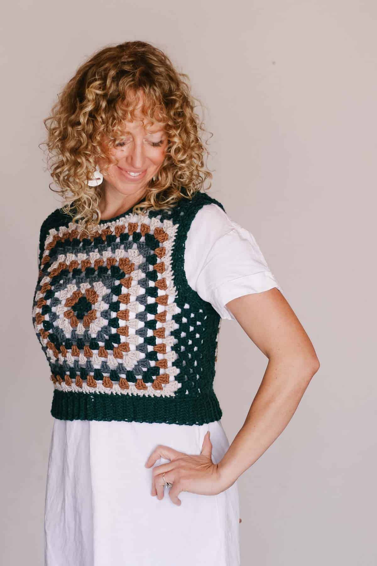 A woman smiling and looking down is wearing a granny square top.