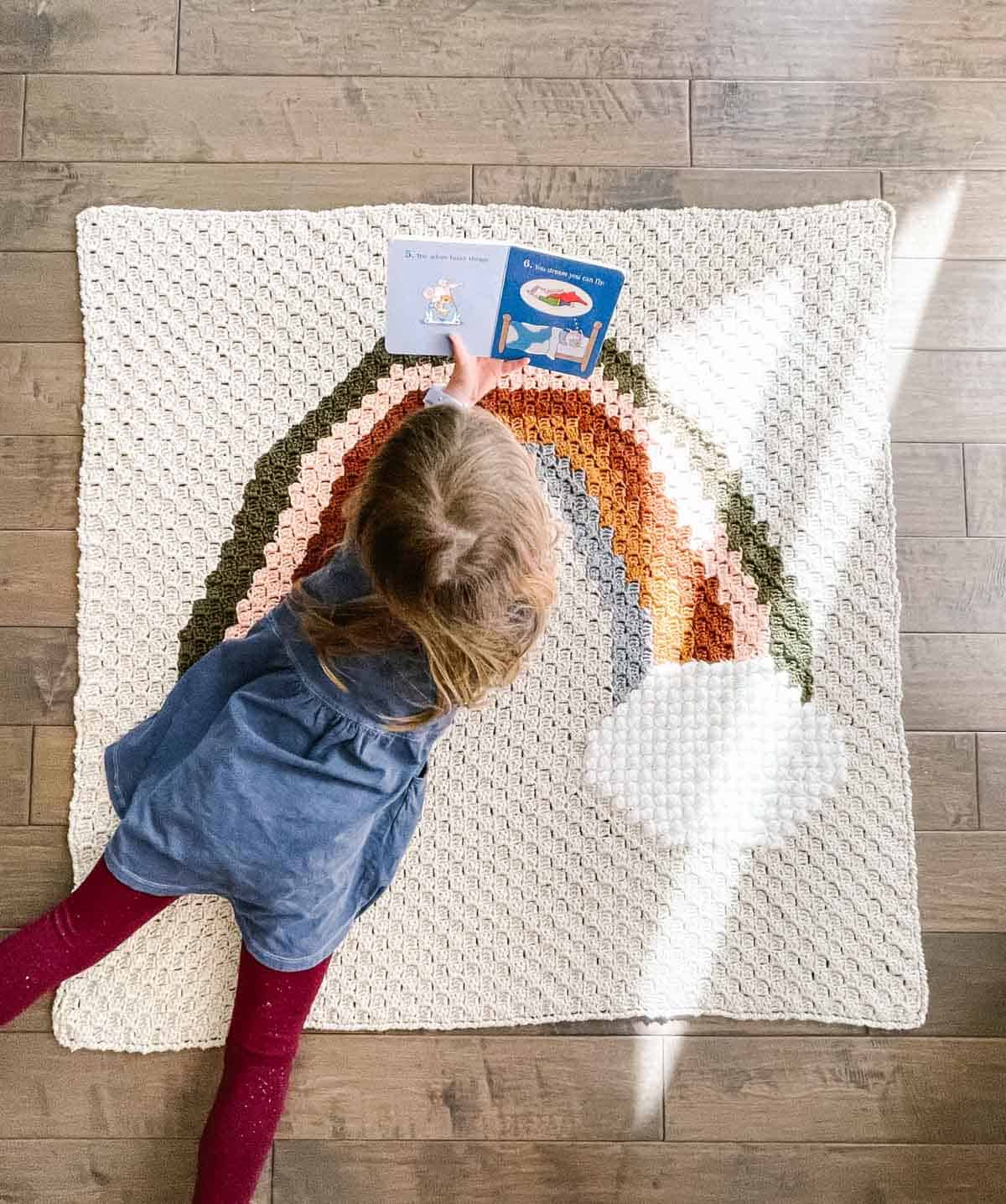 Girl reading while laying on a modern crochet rainbow blanket.