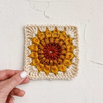 A hand holding a crochet sunburst granny square that looks like a sunflower.