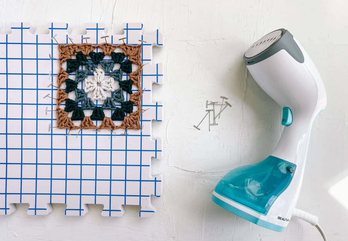 A granny square pinned on a blocking board using T-pins and a steamer.
